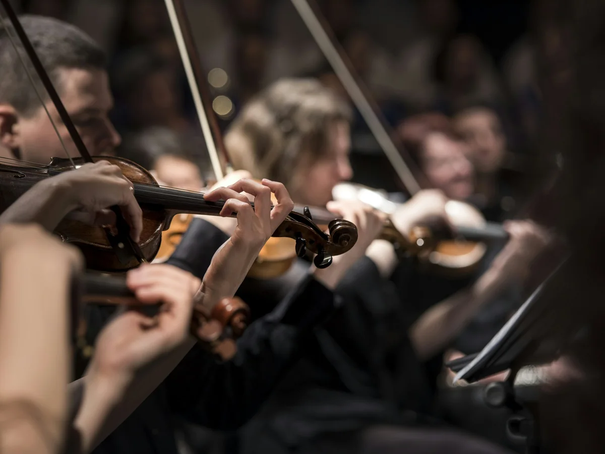 Close-up of string musicians' hands playing violins on a dark stage (representative photograph of a live orchestra, not this show's own musicians)